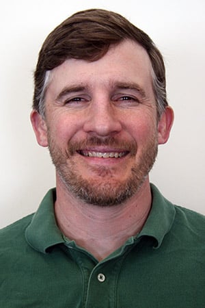 A man with short brown hair and a beard, wearing a green polo shirt, is smiling in front of a plain light background.