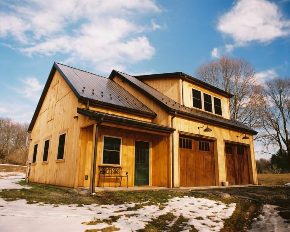 A two-story wooden house with a green door, large garage doors, and a metal roof, surrounded by patchy snow and leafless trees under a blue sky.