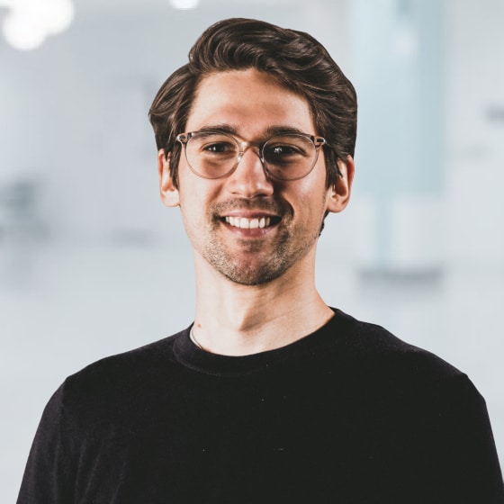 A person with glasses and a beard, wearing a black shirt, smiles in a bright, blurred indoor setting.